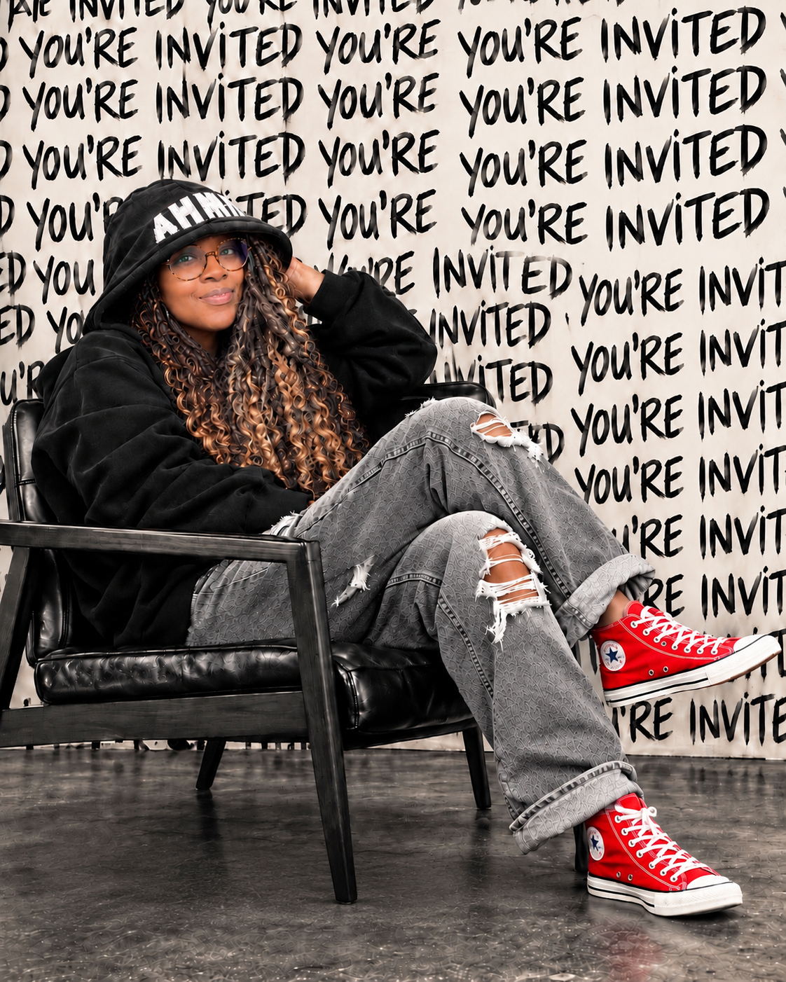 INVITED by Adrienne: woman seated in a black chair wearing a black hoodie, gray jeans, and red Converse sneakers against a handwritten You're Invited backdrop.